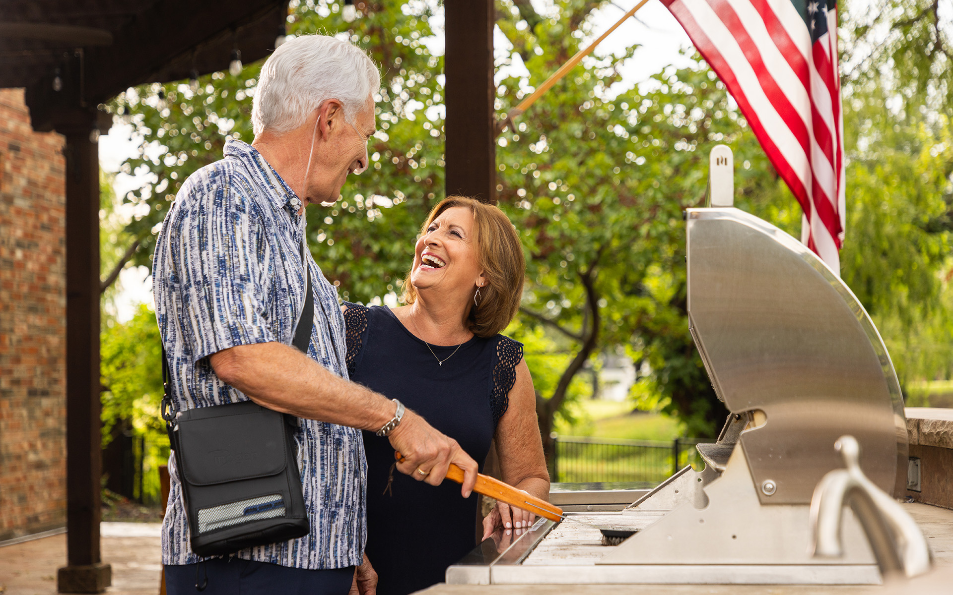A man using an Inogen portable oxygen concentrator while joking with a woman and cleaning his barbecue in his backyard